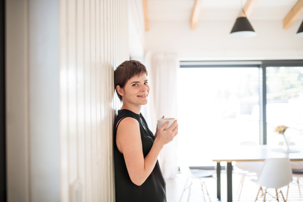 A young woman with coffee standing indoors at home, leaning on wall. Copy space.