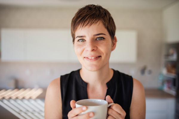 A front view of young woman with coffee standing indoors at home.