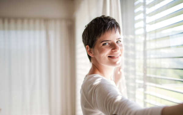 Portrait of young happy woman standing by window indoors at home, looking at camera.