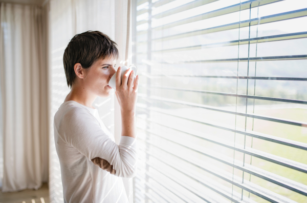 Portrait of young happy woman with coffee standing by window indoors at home, drinking.