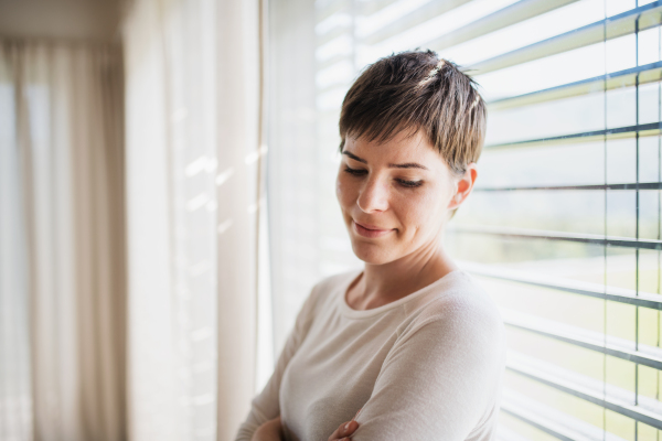 A portrait of young woman standing by window indoors at home, daydreaming.