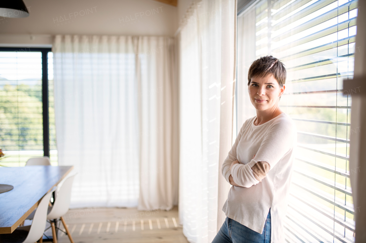 Portrait of young happy woman standing by window indoors at home, looking at camera.