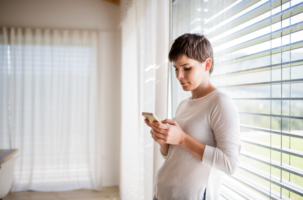 Portrait of young woman with smartphone standing by window indoors at home, text messaging.