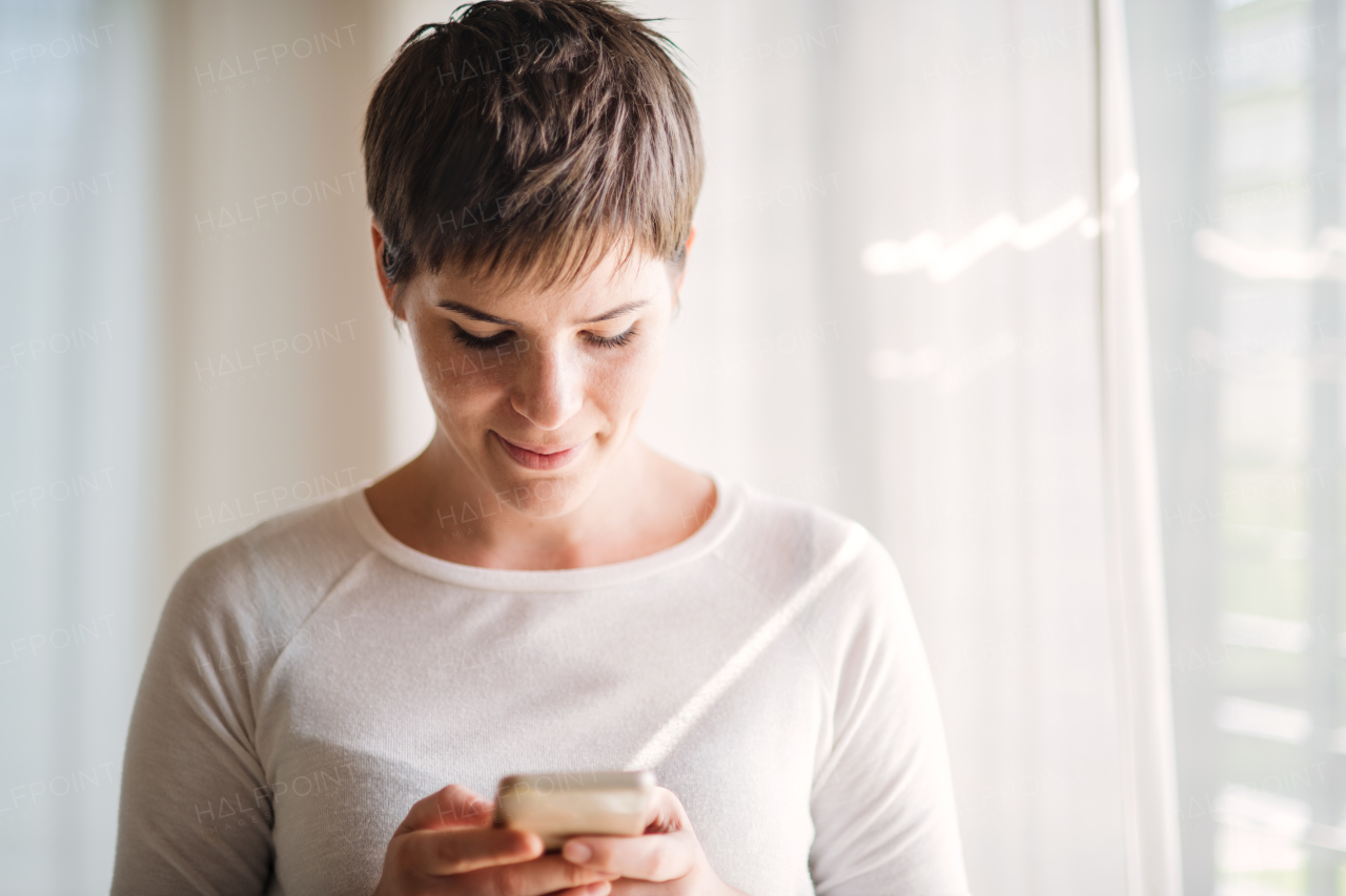 Portrait of young woman with smartphone standing indoors at home, texting.