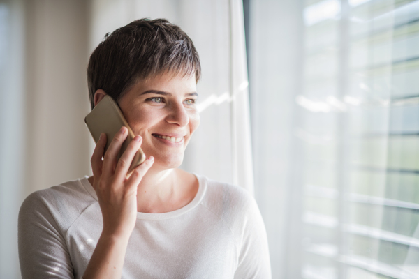 Portrait of young woman with smartphone standing by window indoors at home.