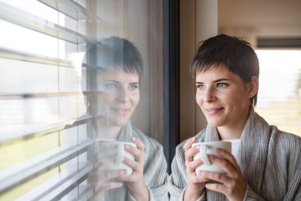 Portrait of young happy woman with coffee standing indoors by window at home.