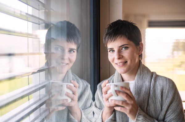 Portrait of young happy woman with coffee standing indoors by window at home.