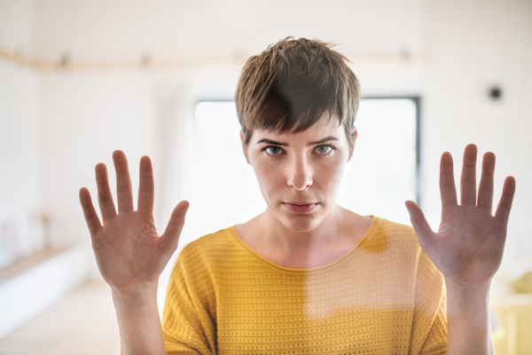 Front view of sad young woman standing indoors at home, looking at camera. Shot through glass.