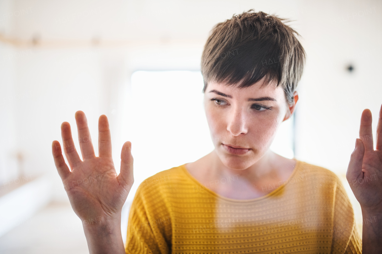 Front view of sad young woman standing indoors at home, hands on window. Shot through glass.