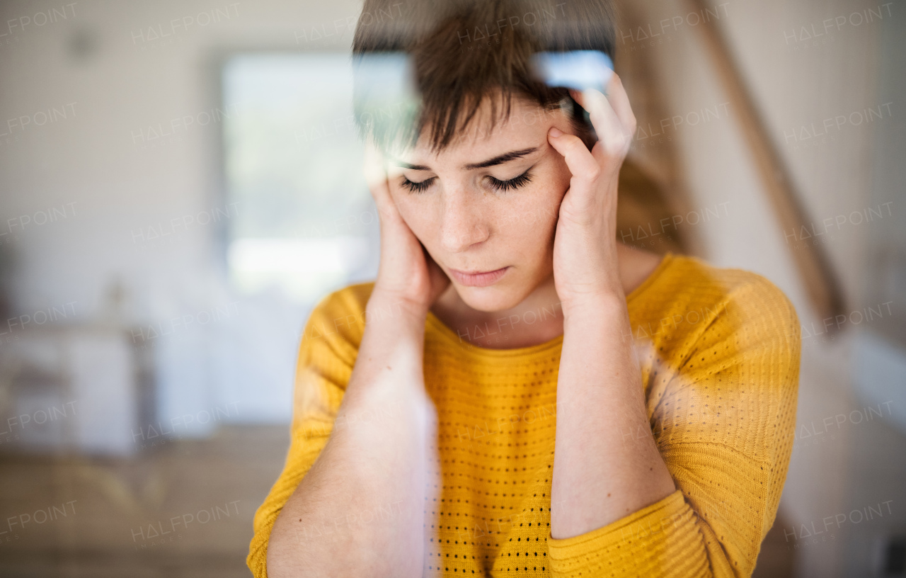 Front view of sad young woman standing indoors at home, close-up. Shot through glass.