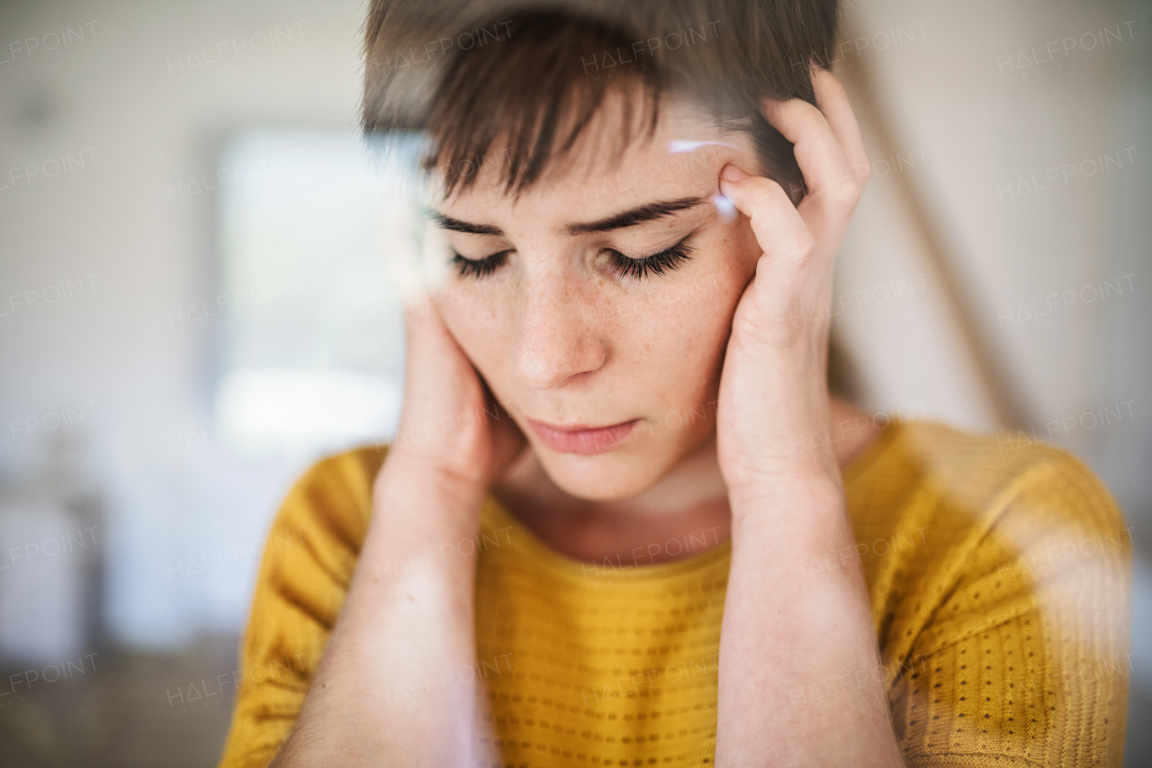 Front view of sad young woman standing indoors at home, close-up. Shot through glass.