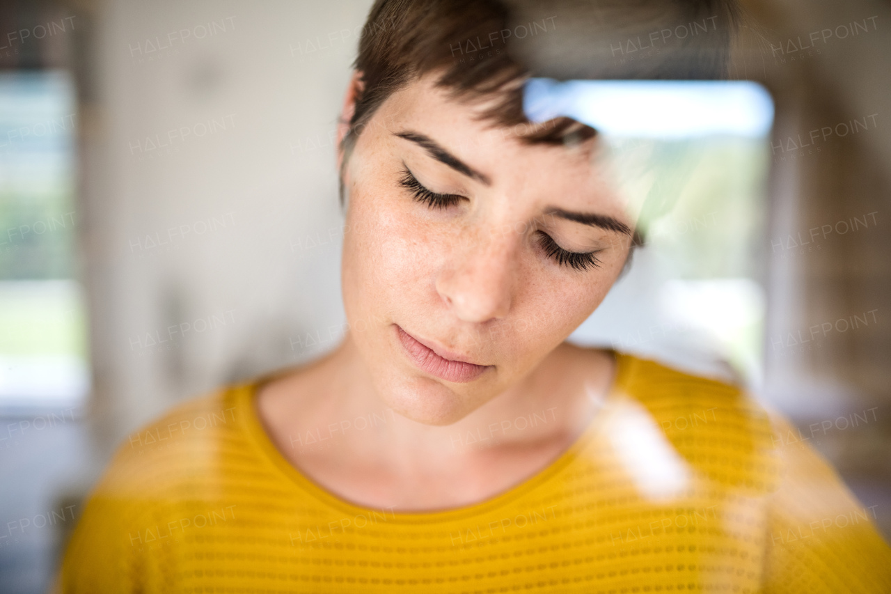 Front view of sad young woman standing indoors at home, close-up. Shot through glass.