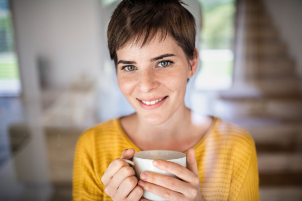 Front view of young woman with coffee standing indoors at home. Shot through glass.