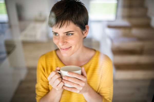 Front view of young woman with coffee standing indoors at home. Shot through glass.