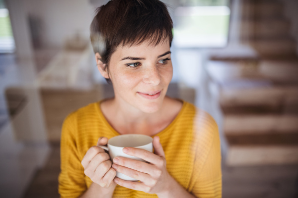 Front view of young woman with coffee standing indoors at home. Shot through glass.