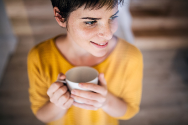 Front view of young woman with coffee standing indoors at home. Shot through glass.