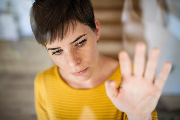 Front view of sad young woman standing indoors at home, close-up. Shot through glass.