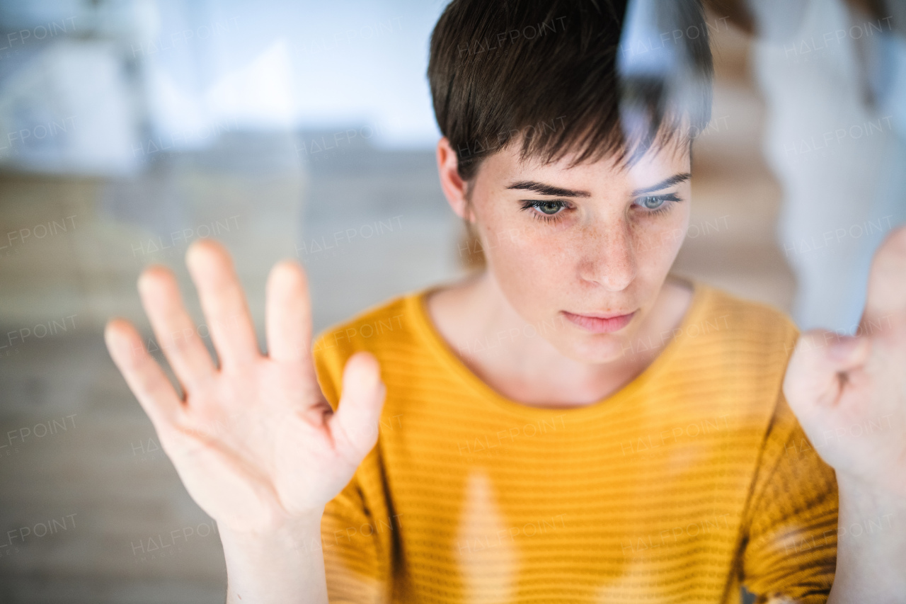 Front view of sad young woman standing indoors at home, looking at camera. Shot through glass.