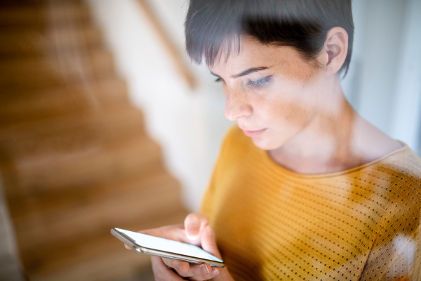 A young woman with smartphone standing indoors at home. Shot through glass.