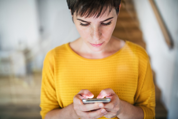 Front view of young woman with smartphone standing indoors at home. Shot through glass.