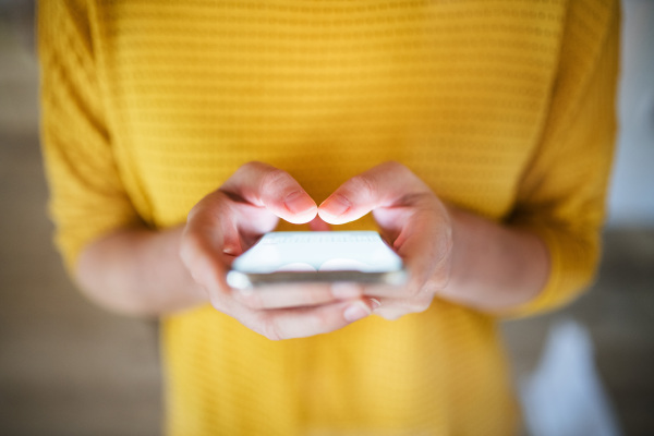 Midsection of young woman with smartphone standing indoors at home. Shot through glass.