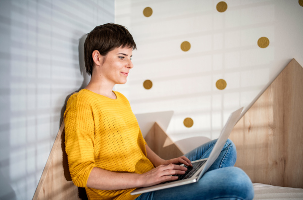 Side view of young woman with laptop sitting on bed in bedroom indoors at home.