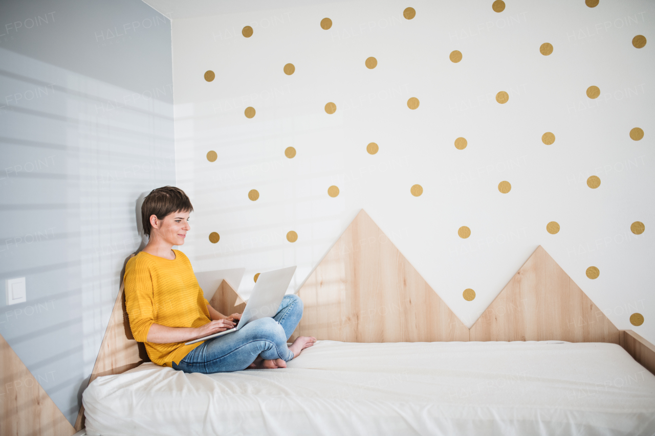 Side view of young woman with laptop sitting on bed in bedroom indoors at home.