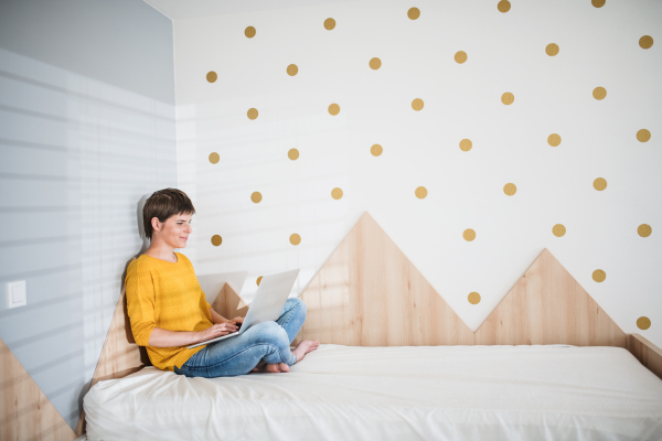 Side view of young woman with laptop sitting on bed in bedroom indoors at home.