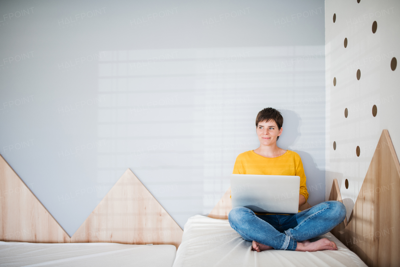 Front view of young woman with laptop sitting on bed in bedroom indoors at home. Copy space.