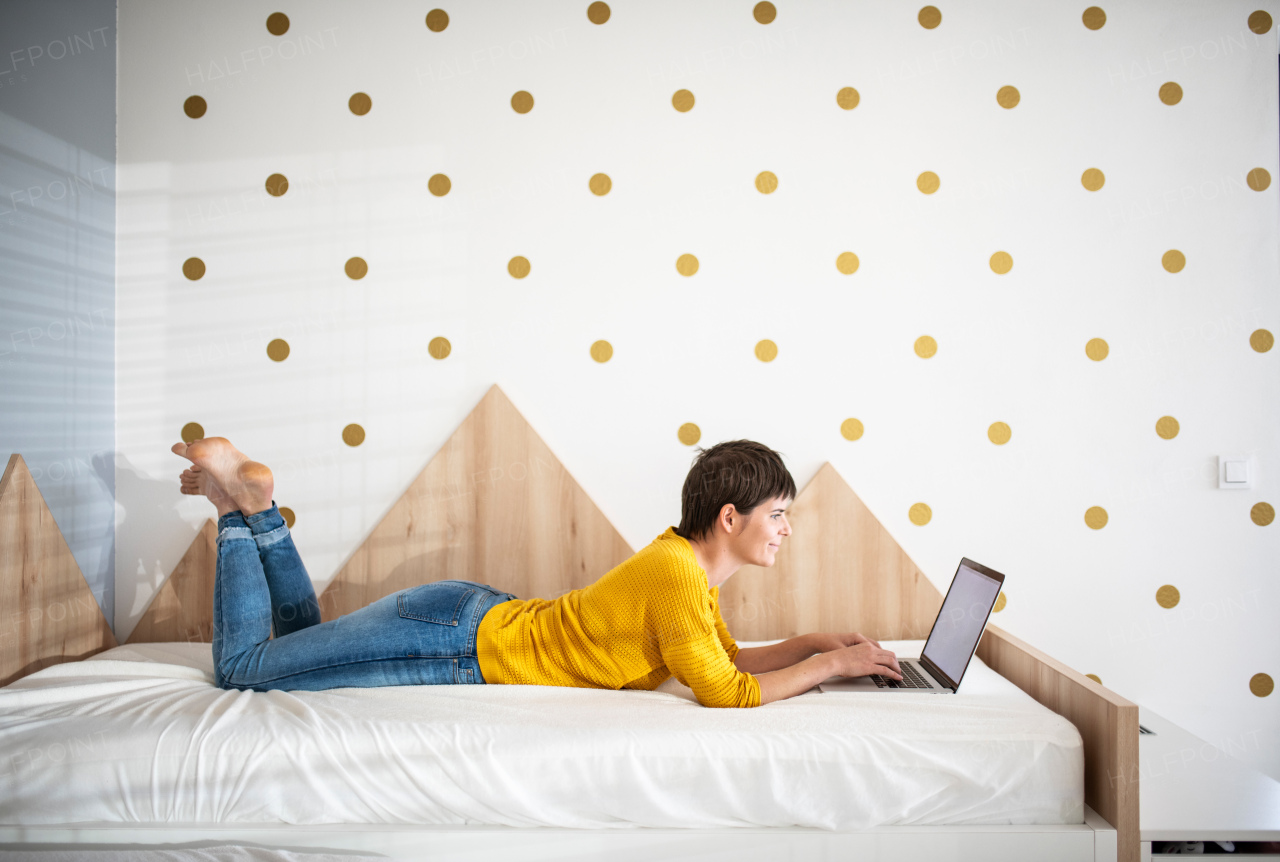 Side view of young woman with laptop lying on bed in bedroom indoors at home.