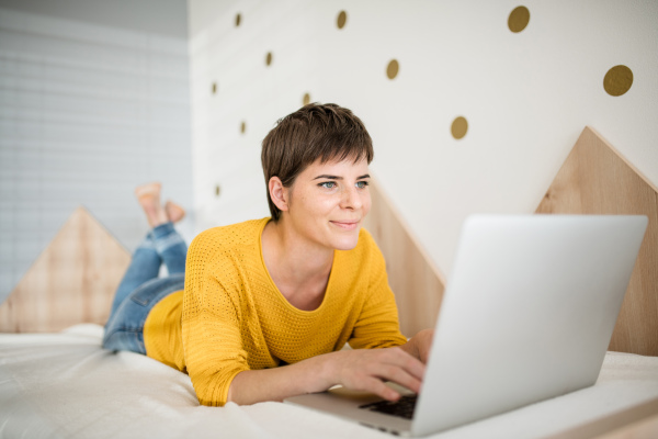 A young woman with laptop lying on bed in bedroom indoors at home.