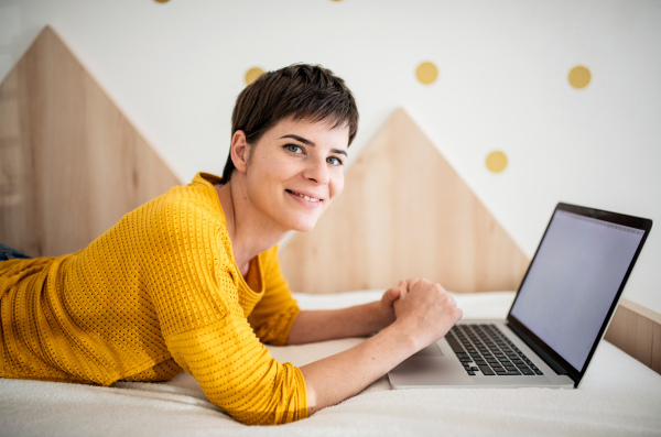 Side view of young woman with laptop lying on bed in bedroom indoors at home.