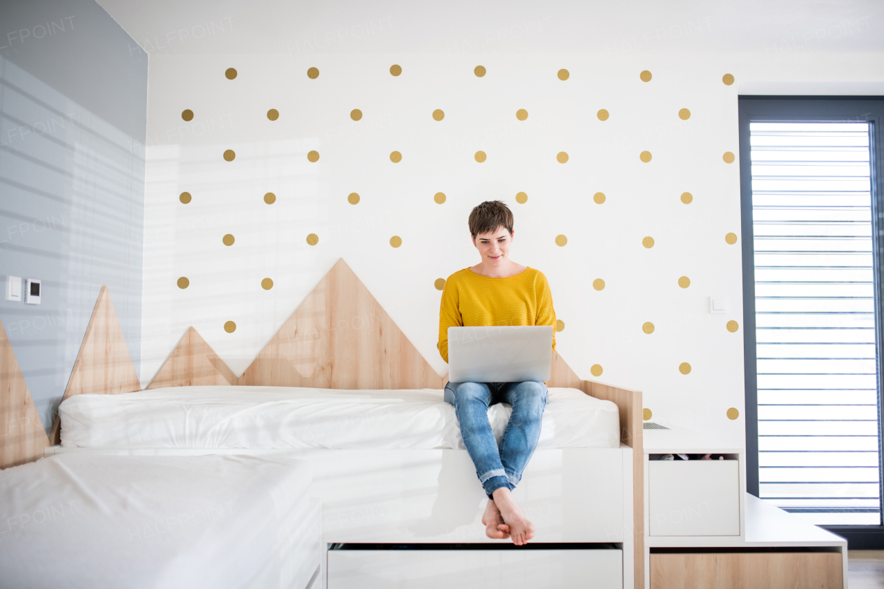 Front view of young woman with laptop sitting on bed in bedroom indoors at home.