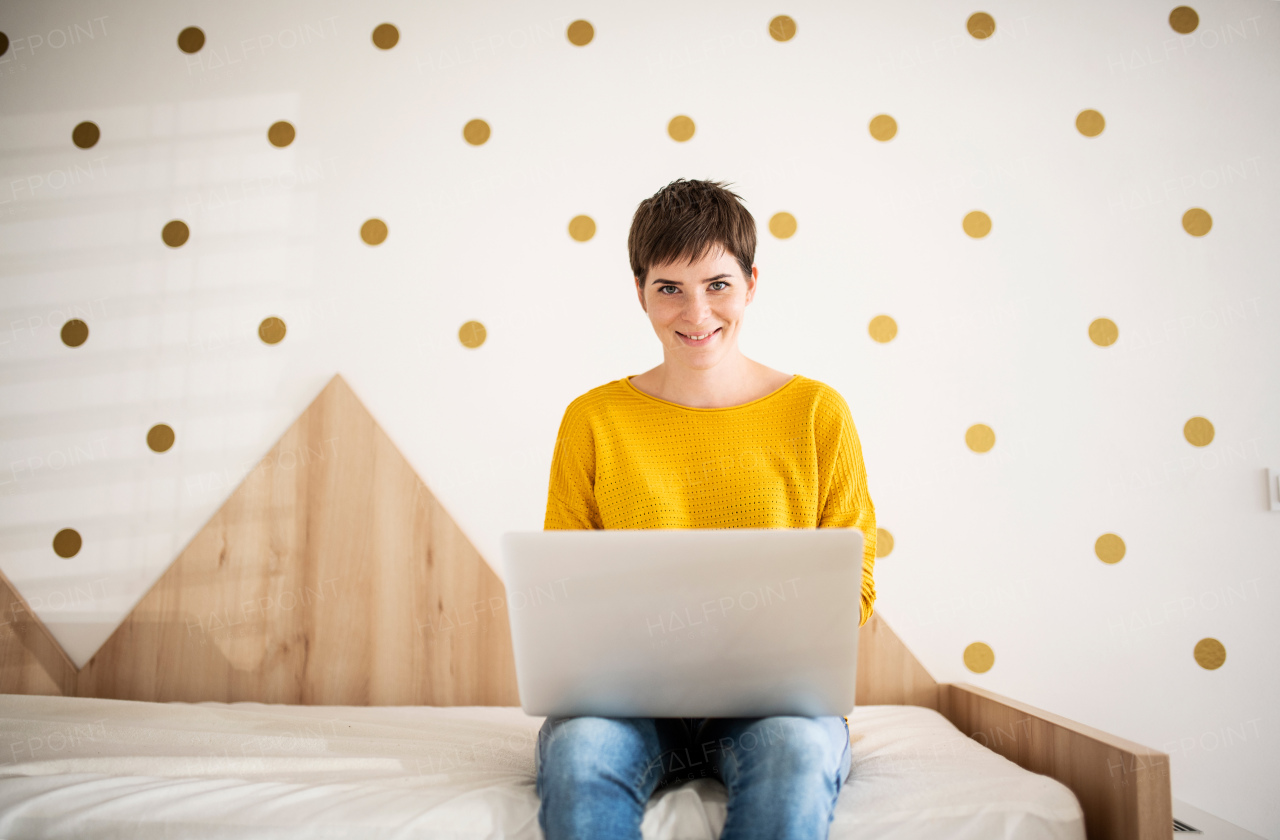 Front view of young woman with laptop sitting on bed in bedroom indoors at home.