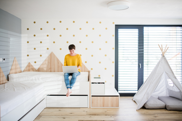 Front view of young woman with laptop sitting on bed in bedroom indoors at home.