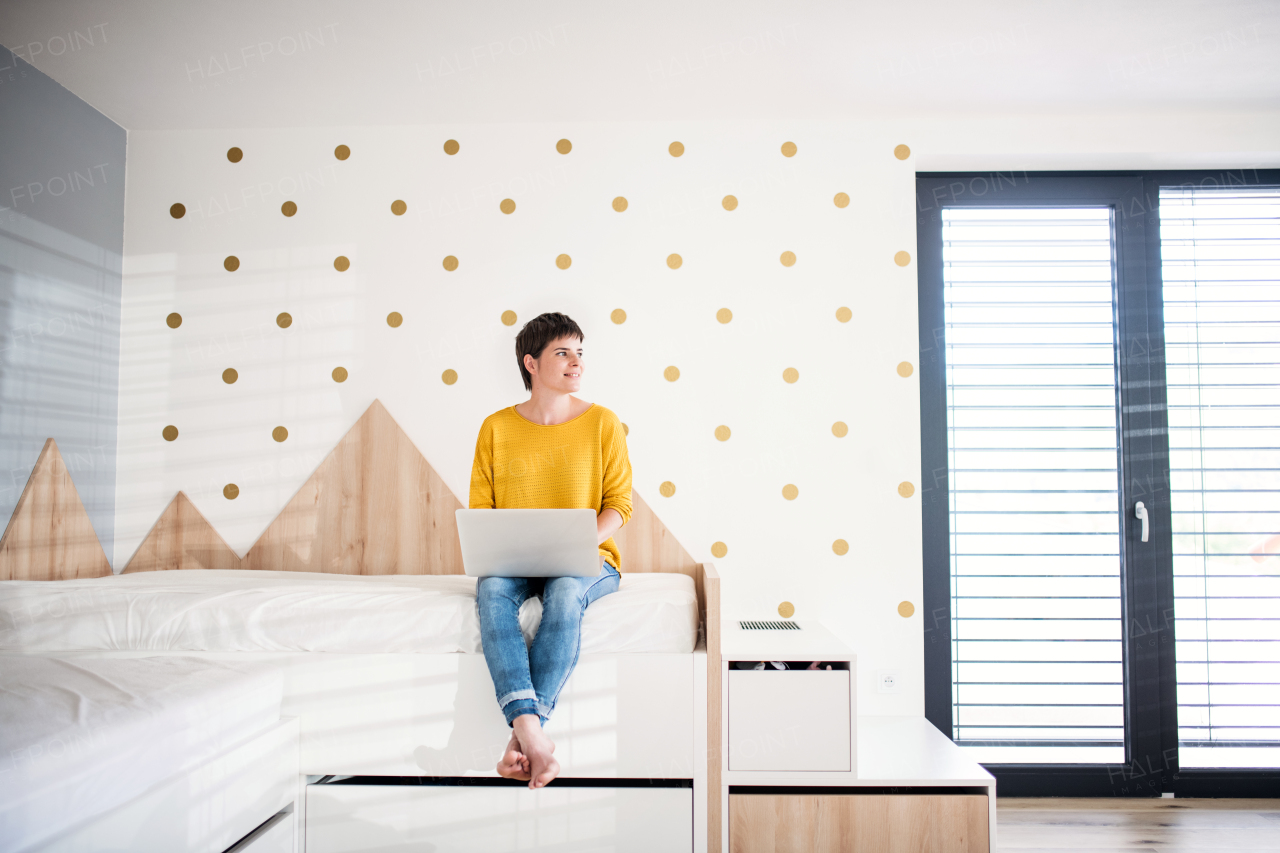 Front view of young woman with laptop sitting on bed in bedroom indoors at home.