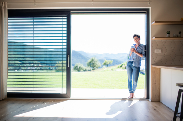 Front view of young woman with coffee standing by patio door at home. Copy space.