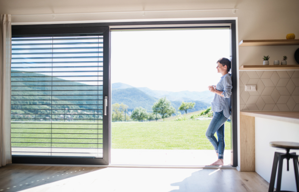 Side view of young woman with coffee standing by patio door at home. Copy space.