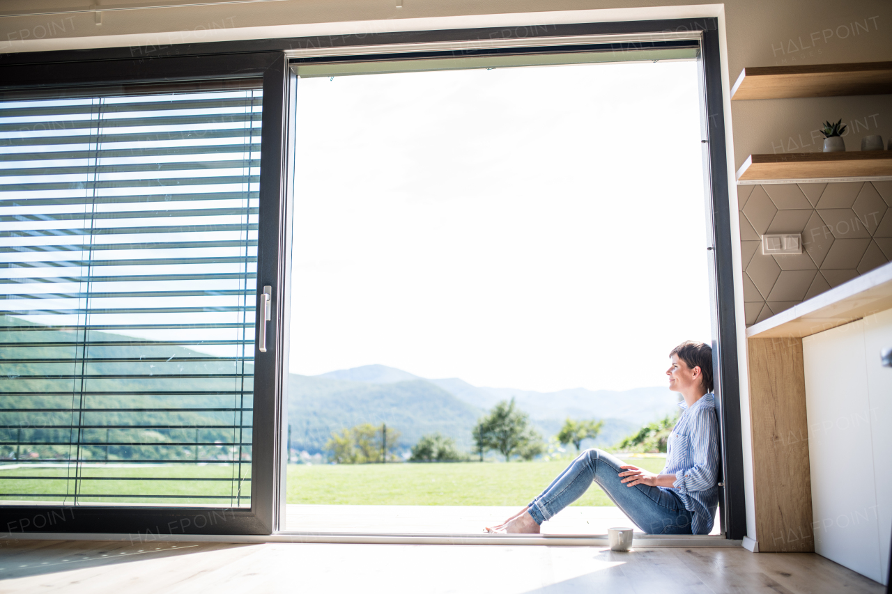 Side view of young woman with coffee sitting by patio door at home. Copy space.