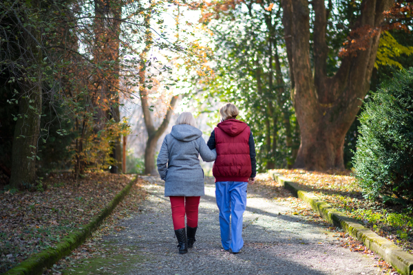 A rear view of senior woman with caregiver outdoors on a walk in park, coronavirus concept.