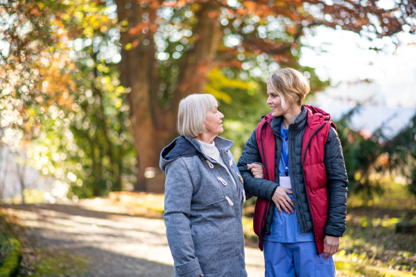 Senior woman with caregiver or healthcare worker outdoors on a walk in park, coronavirus concept.