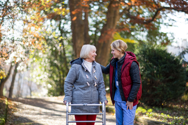 Senior woman with walking frame and caregiver or healthcare worker outdoors on a walk in park, coronavirus concept.