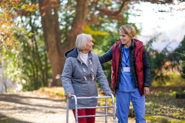 Senior woman with walking frame and caregiver or healthcare worker outdoors on a walk in park.