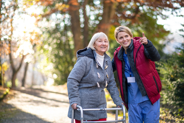 Senior woman with walking frame and caregiver or healthcare worker outdoors on a walk in park, coronavirus concept.