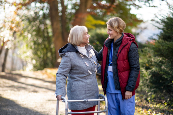 Senior woman with walking frame and caregiver or healthcare worker outdoors on a walk in park, coronavirus concept.