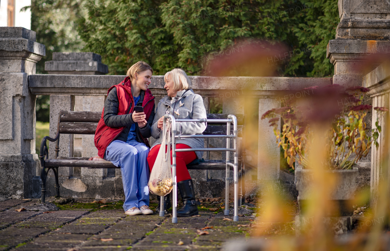 Senior woman with walking frame and caregiver outdoors sitting on bench in park, coronavirus concept.