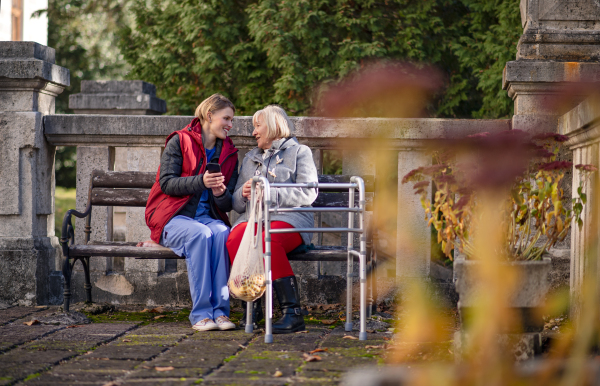 Senior woman with walking frame and caregiver outdoors sitting on bench in park, coronavirus concept.