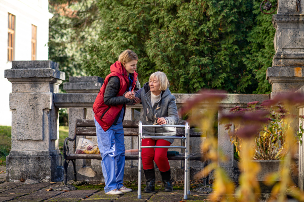 Senior woman with walking frame and caregiver outdoors sitting on bench in park, helping to stand up.