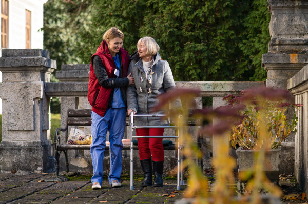 Senior woman with walking frame and caregiver or healthcare worker outdoors on a walk in park.