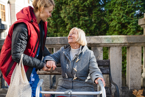 Senior woman with walking frame and caregiver outdoors sitting on bench in park, helping to stand up.